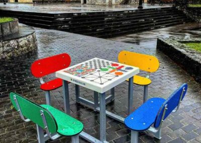 Mesa con juegos de mesa y bancos con respaldo en una plaza urbana.