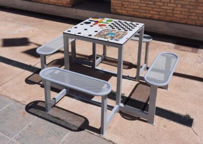 Table avec jeux de société et bancs en acier dans un espace public.